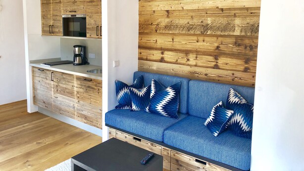 Un espace de vie compact comprenant un coin cuisine, un canapé bleu orné de coussins à motifs et un mur en bois.