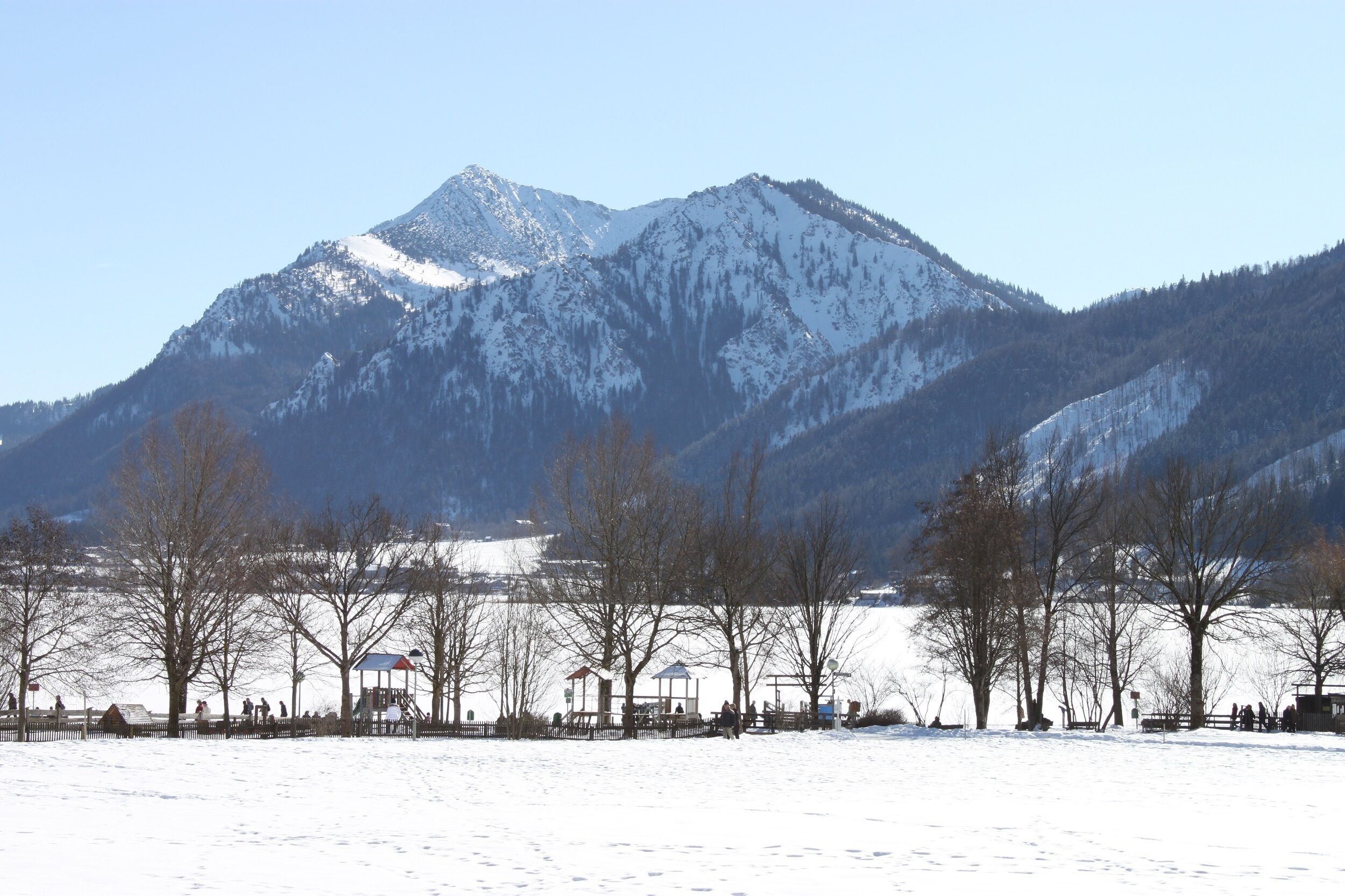 A snow-covered park with a playground and benches, surrounded by mountains.