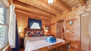Camera da letto in una baita di legno, con un letto, un comodino, una lampada, una finestra con tende e una porta.