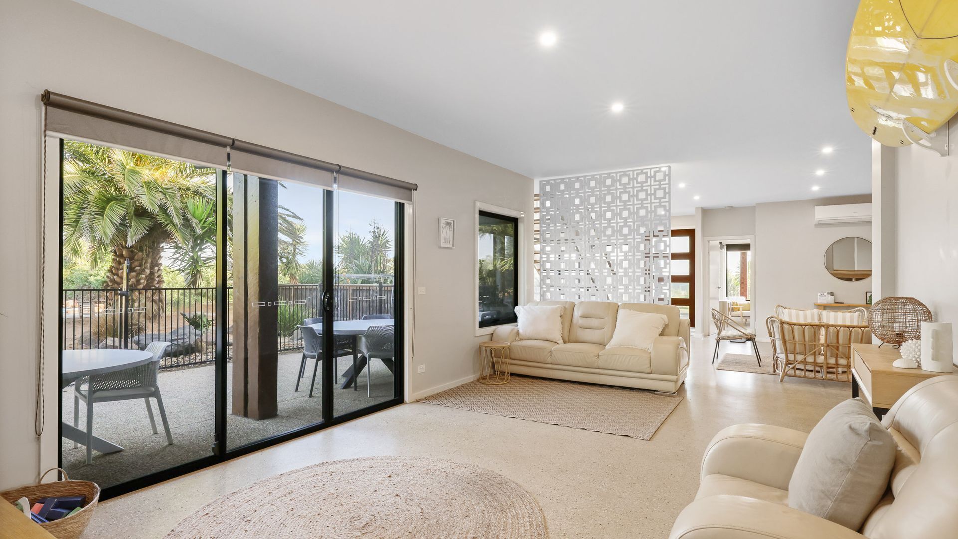 A modern living room with a sliding glass door leading to an outdoor patio, a beige sofa, a dining area with wicker chairs, and a decorative wall panel.