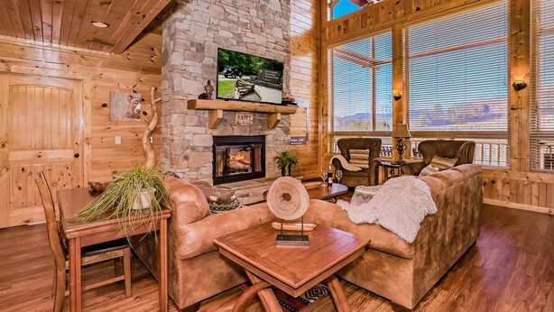 Interior acogedor de una cabaña con chimenea de piedra, muebles de madera y ventanas amplias que ofrecen una vista.