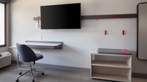 Un espacio de oficina moderno con una televisión de pantalla plana montada en la pared, un escritorio con una silla y un estante pequeño con un cojín.