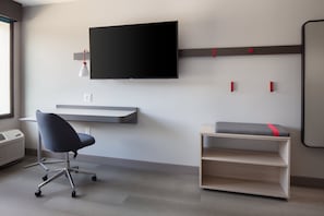 Un espacio de oficina moderno con una televisión de pantalla plana montada en la pared, un escritorio con una silla y un estante pequeño con un cojín.
