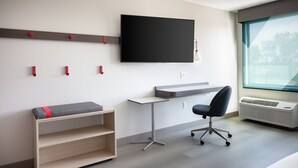 Una habitación moderna con un televisor de pantalla plana en la pared, un escritorio con silla, un banco con espacio de almacenamiento y un amplio ventanal que proporciona luz natural.