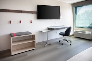 Una habitación moderna con un televisor de pantalla plana en la pared, un escritorio con silla, un banco con espacio de almacenamiento y un amplio ventanal que proporciona luz natural.