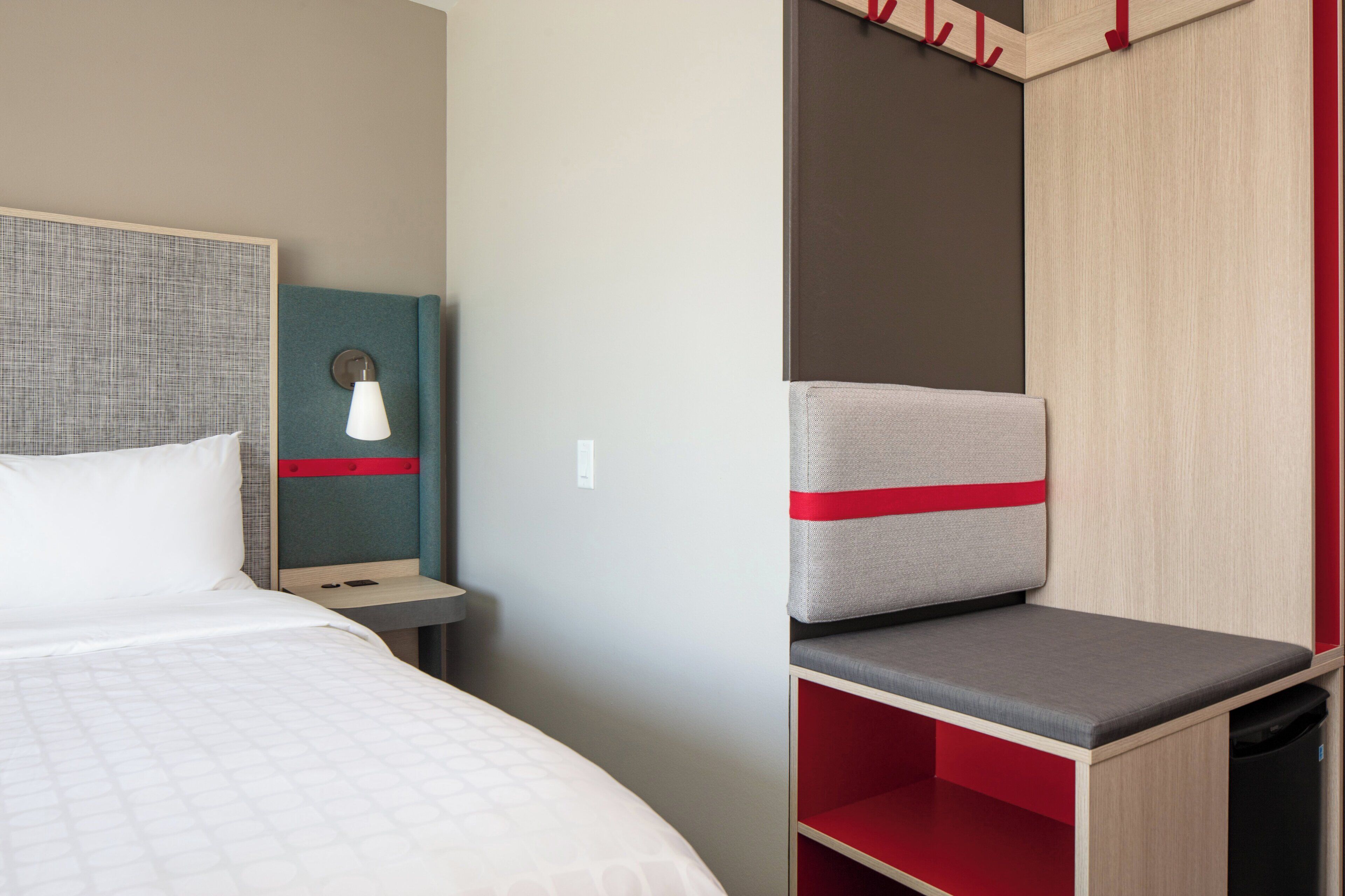 A hotel room with a bed, a bedside table, a lamp, and a seating area with a red cushion.