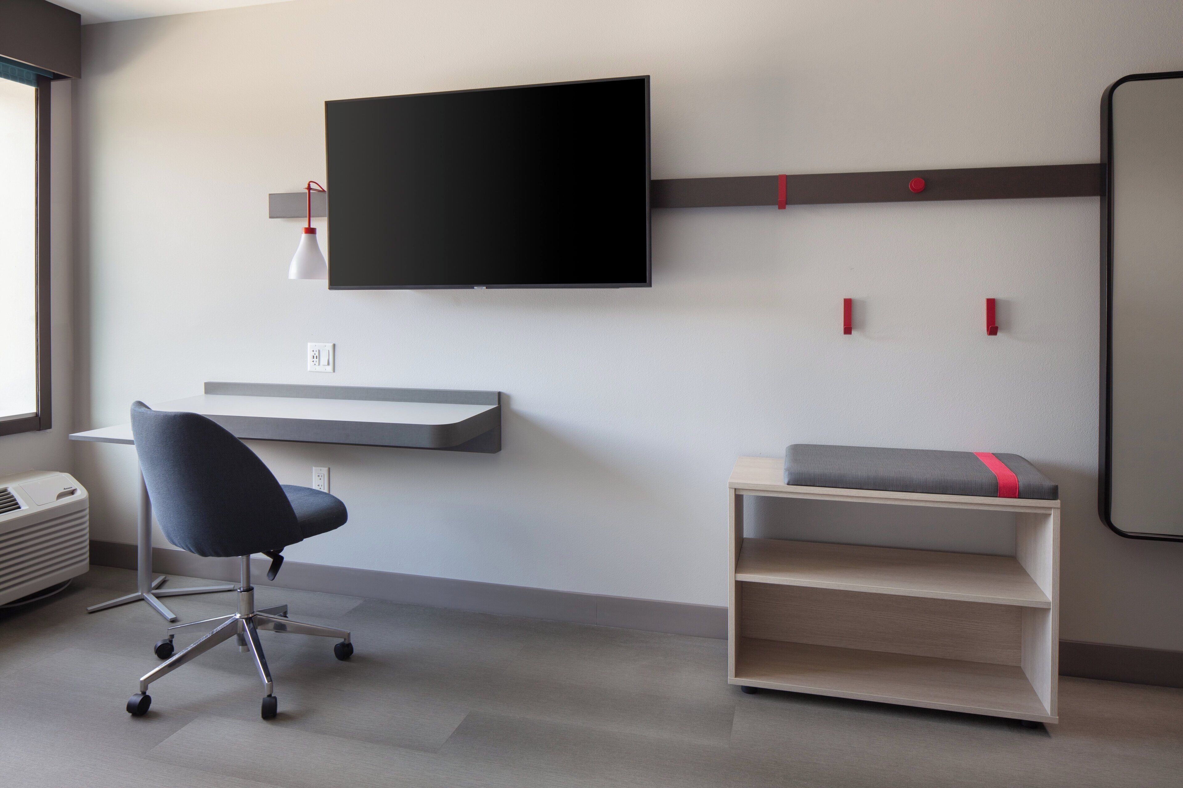 Een moderne kamer met een flatscreen televisie aan de muur, een bureau met stoel, een kleine bank met kussen, en een verwarming op de vloer.