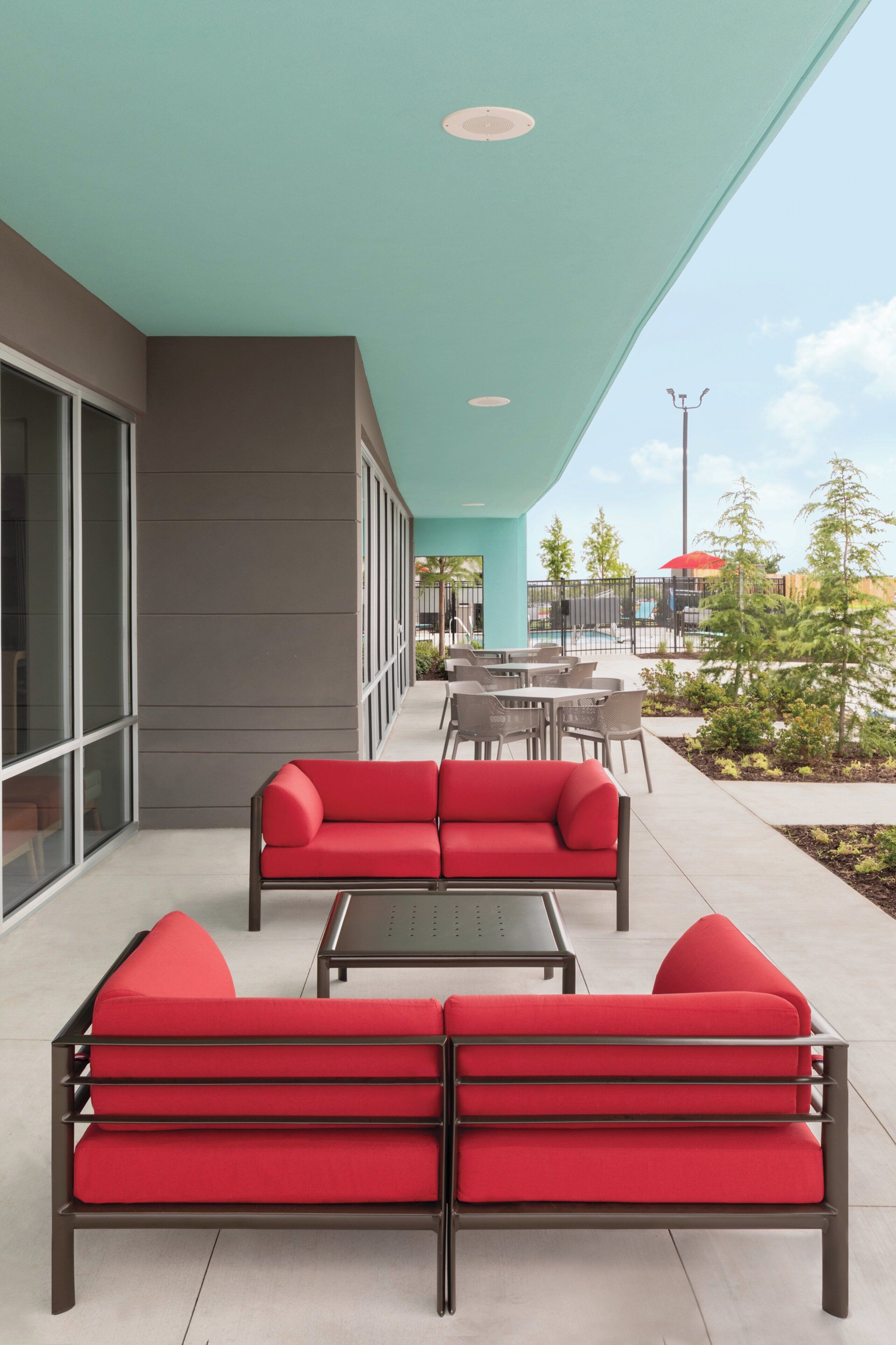 An outdoor seating area with red sofas and a table, a pool in the background, and a building with large windows.