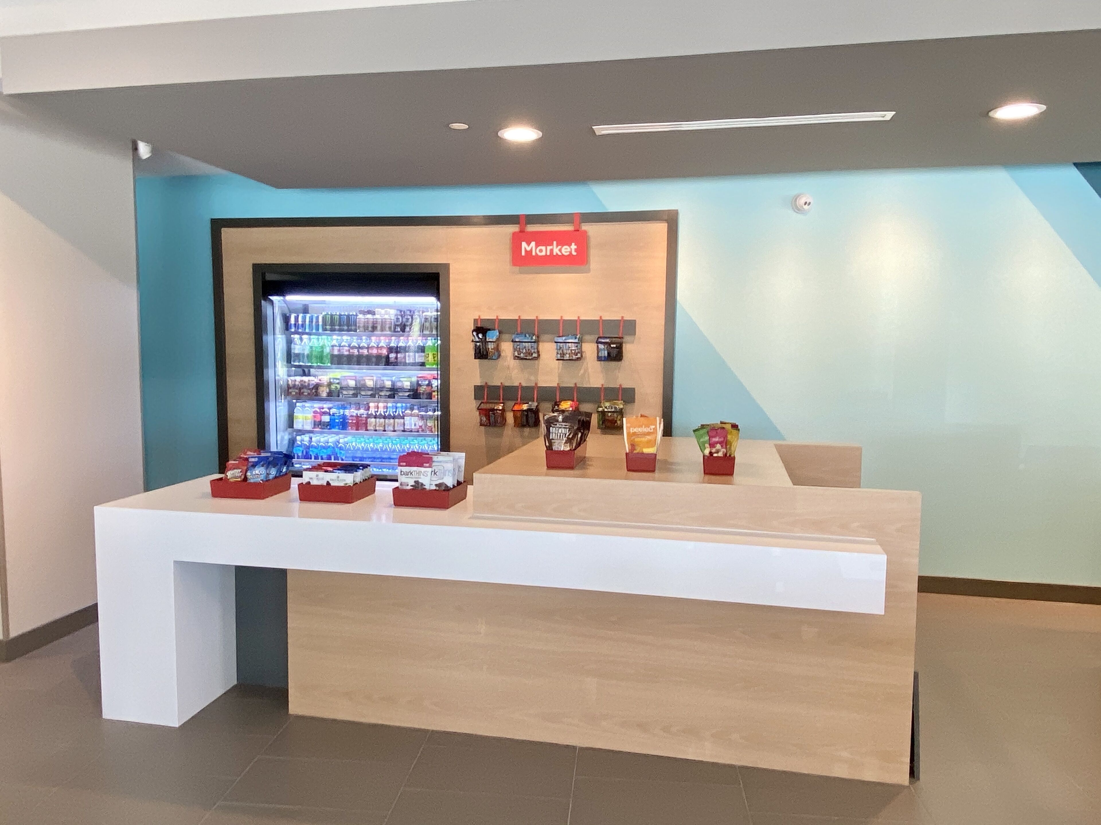 A clean, modern kiosk with a 'Market' sign, a refrigerated beverage display, and various packaged goods.