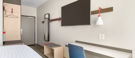 A modern hotel room with a bed, a desk, a chair, a TV mounted on the wall, and a mirror.
