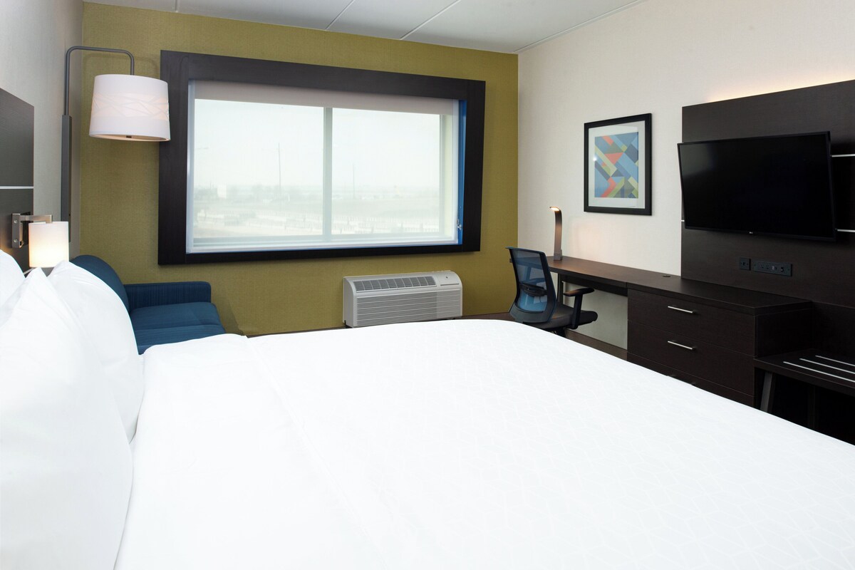 Een hotelkamer met een bed, een bureau, een stoel, een televisie, een raam en een wandairconditioner.