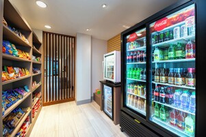 Espaço com petiscos e bebidas, com prateleiras cheias de diversos snacks e um refrigerador exposto com bebidas.