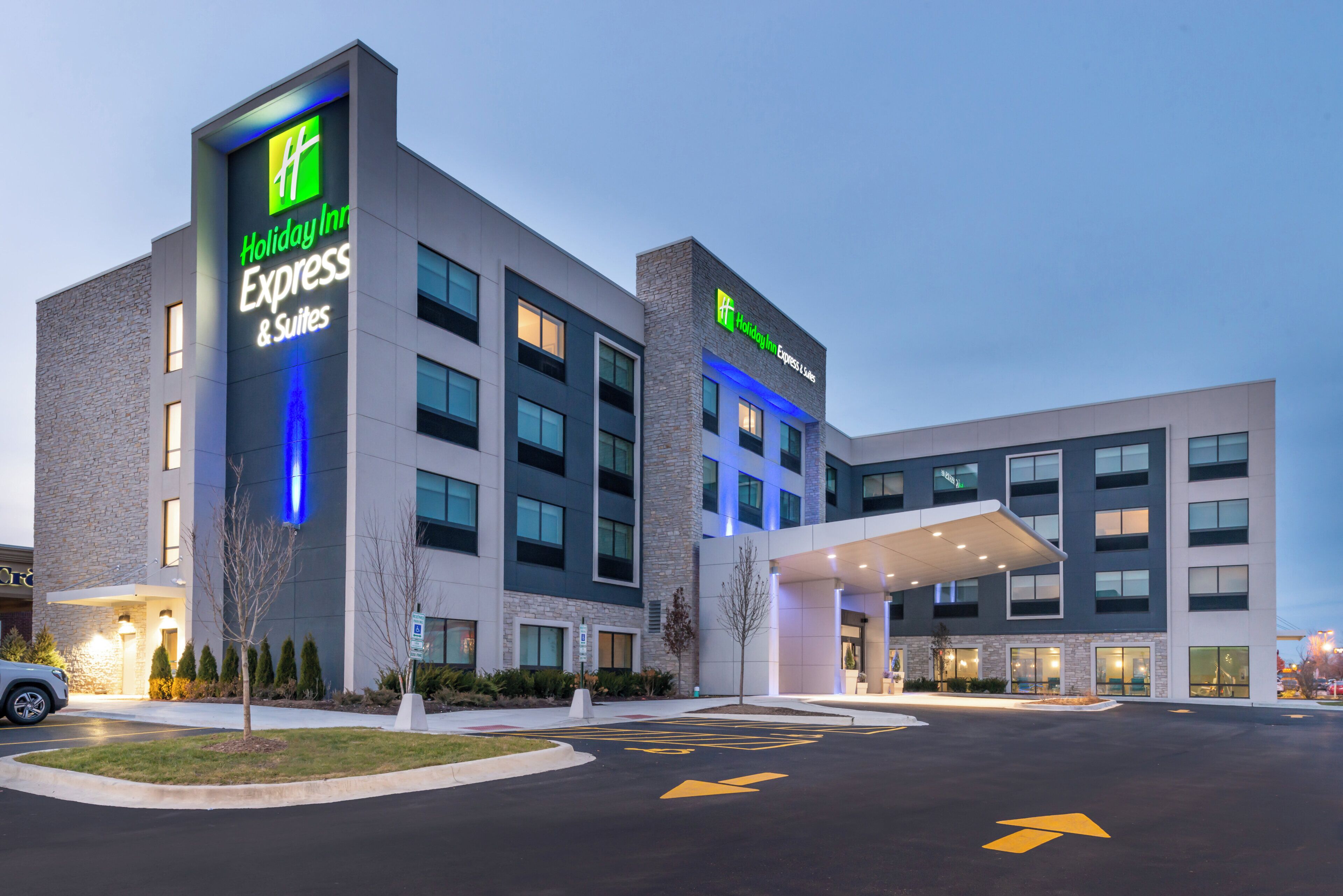 Un hotel Holiday Inn Express & Suites con un diseño moderno, que cuenta con un letrero llamativo, una entrada techada y un estacionamiento con flechas indicativas.