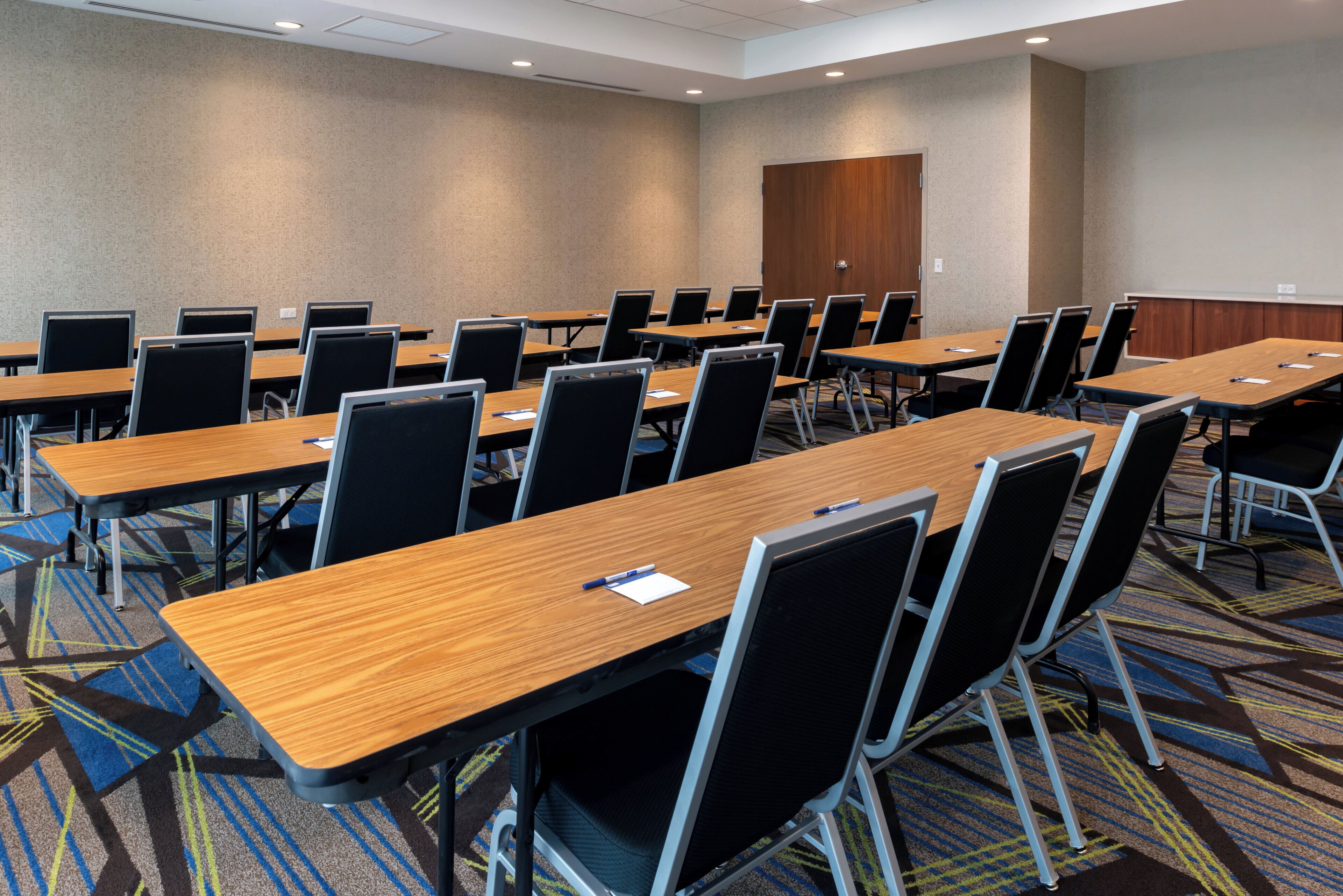 Un salón de conferencias con varias mesas y sillas, piso alfombrado y una puerta al fondo.