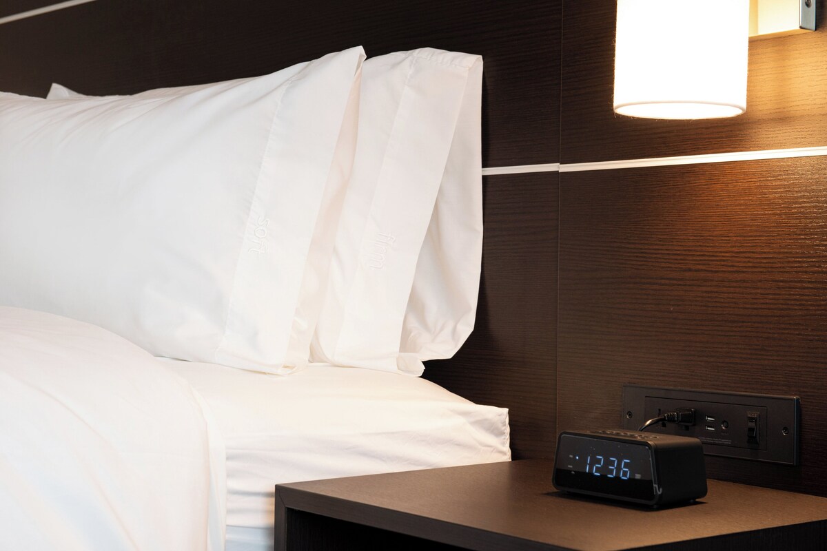 Een net opgemaakt bed met witte lakens, een digitale klok waarop 12:36 staat en een wandlamp.