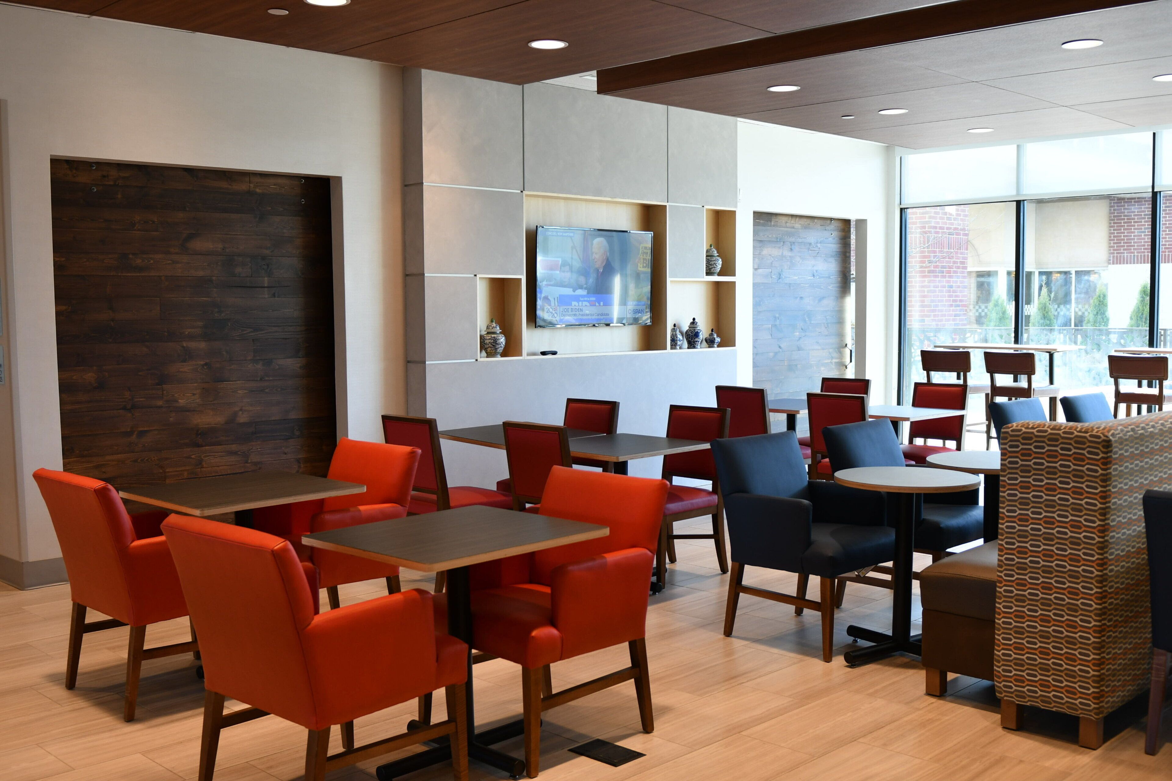 A modern dining area with red and blue chairs, wooden tables, and a large window providing a view of the outside.