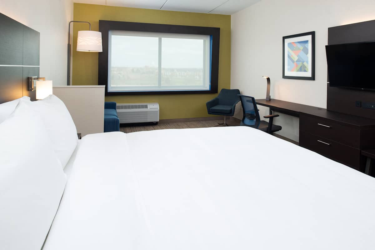 A hotel room with a large bed, a desk with a chair, a flat-screen TV, a window with a view, and a wall-mounted lamp.