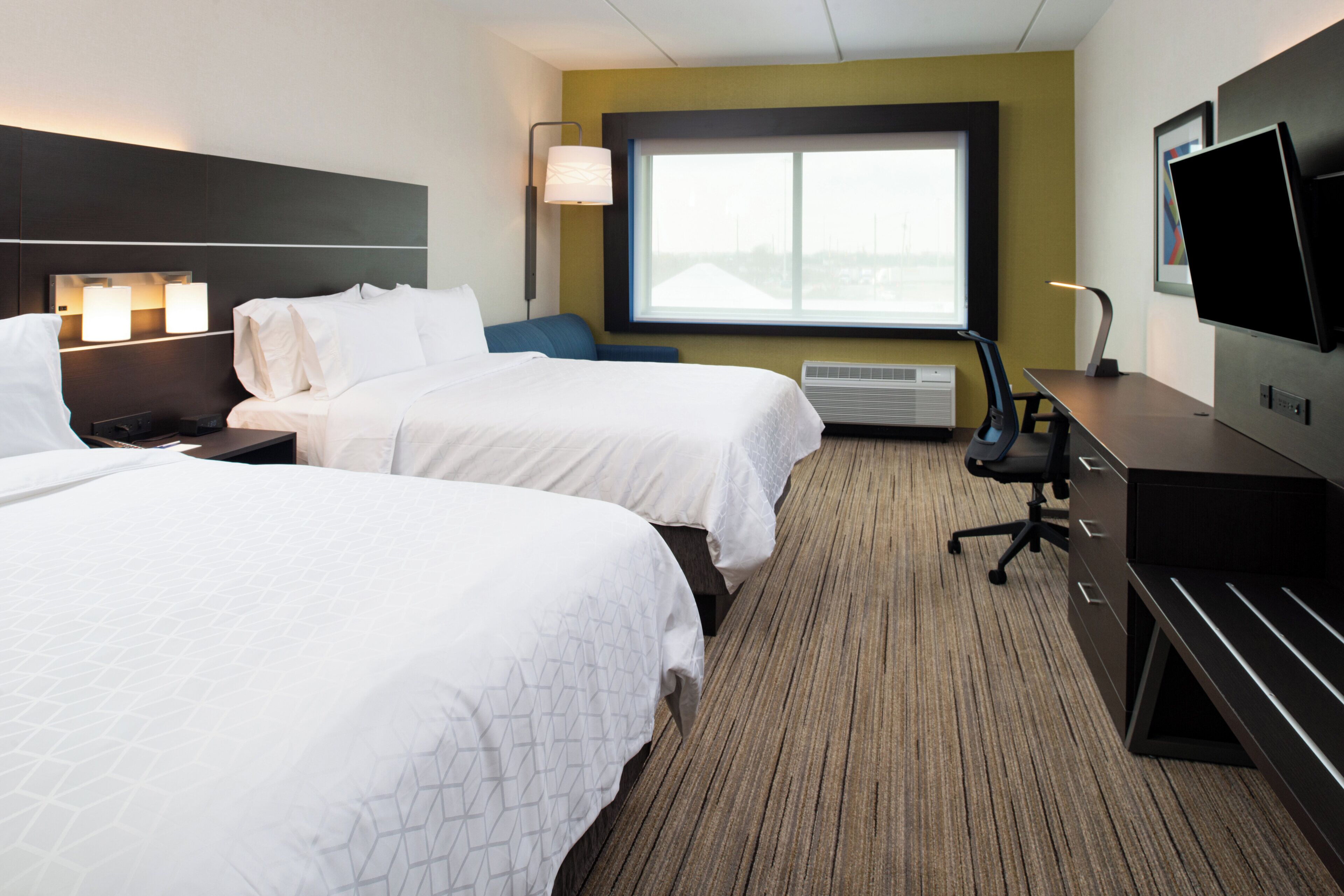 A hotel room with two beds, a desk, a chair, a TV, and a window.