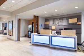 A hotel reception area with a modern design, featuring a reception desk with illuminated panels, a wall-mounted sign for Holiday Inn Express, and a refrigerator in the background. - Holiday Inn Express & Suites Romeoville - Joliet North by IHG (Romeoville)