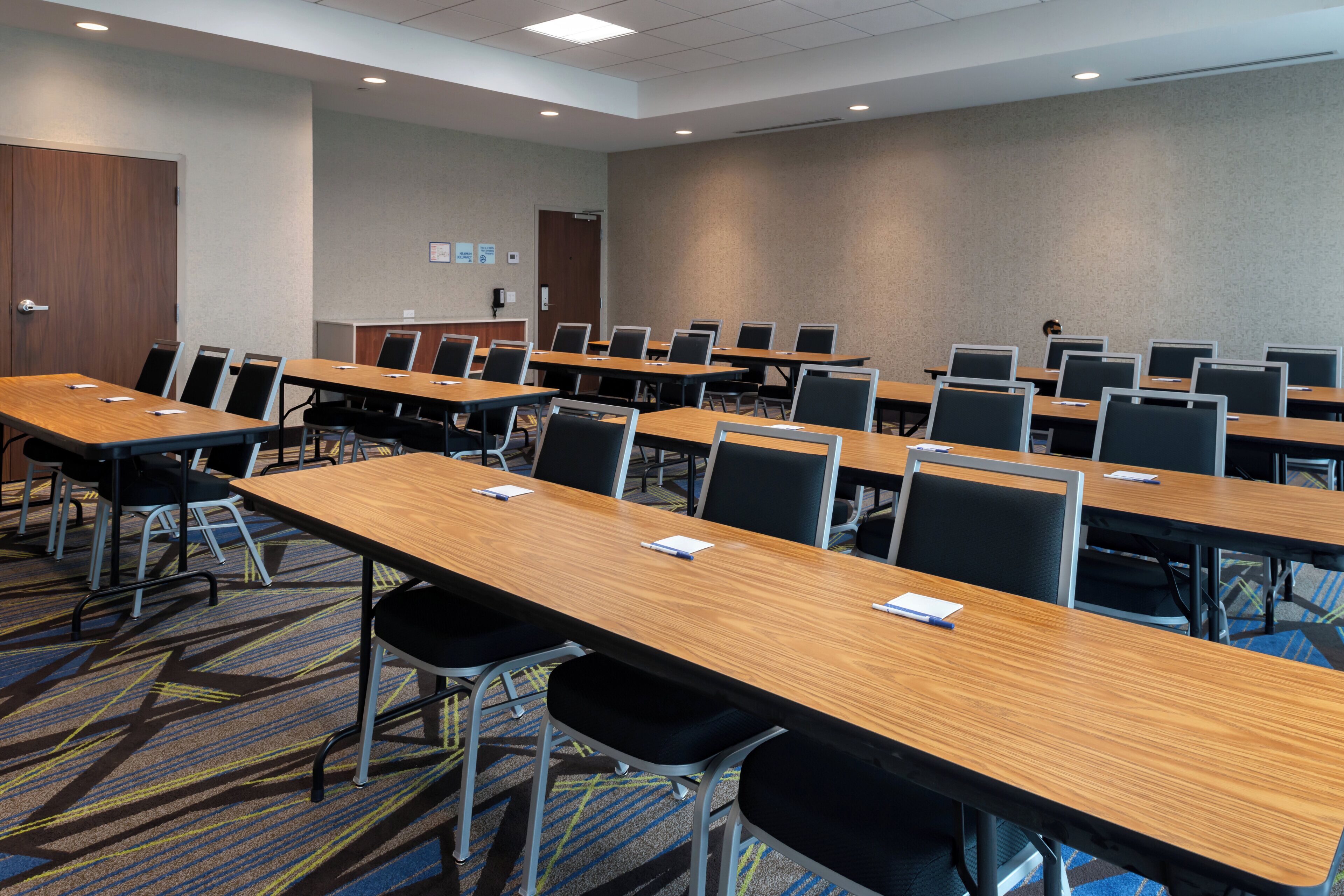 A conference room with multiple tables and chairs, carpeted flooring, and a plain wall with a door and a light switch.