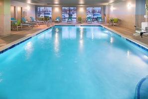 Una amplia piscina interior con agua cristalina y azul, rodeada de sillones de descanso y vista al estacionamiento.