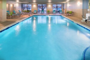 Uma grande piscina interior com água azul cristalina, rodeada por espreguiçadeiras e vista para o estacionamento.