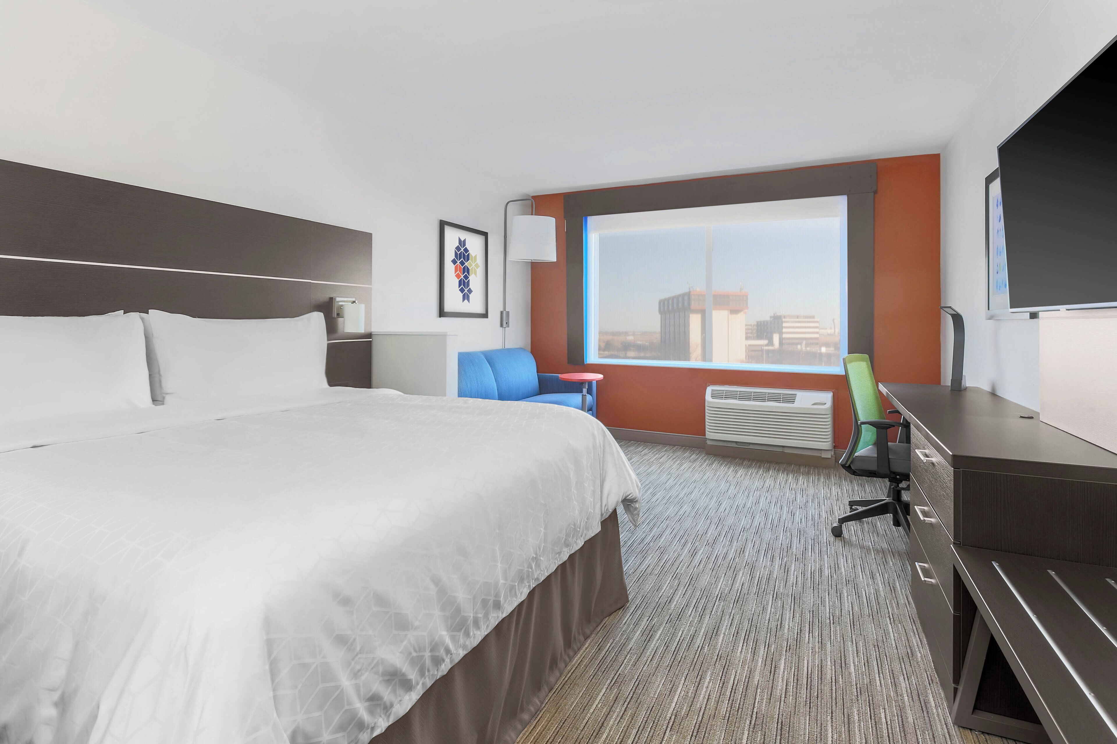 A hotel room with a large bed, a desk, a chair, a window with a city view, and an air conditioning unit.