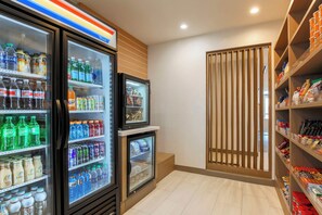 Un refrigerador lleno de bebidas variadas, un refrigerador más pequeño debajo, y estantes con aperitivos y condimentos.