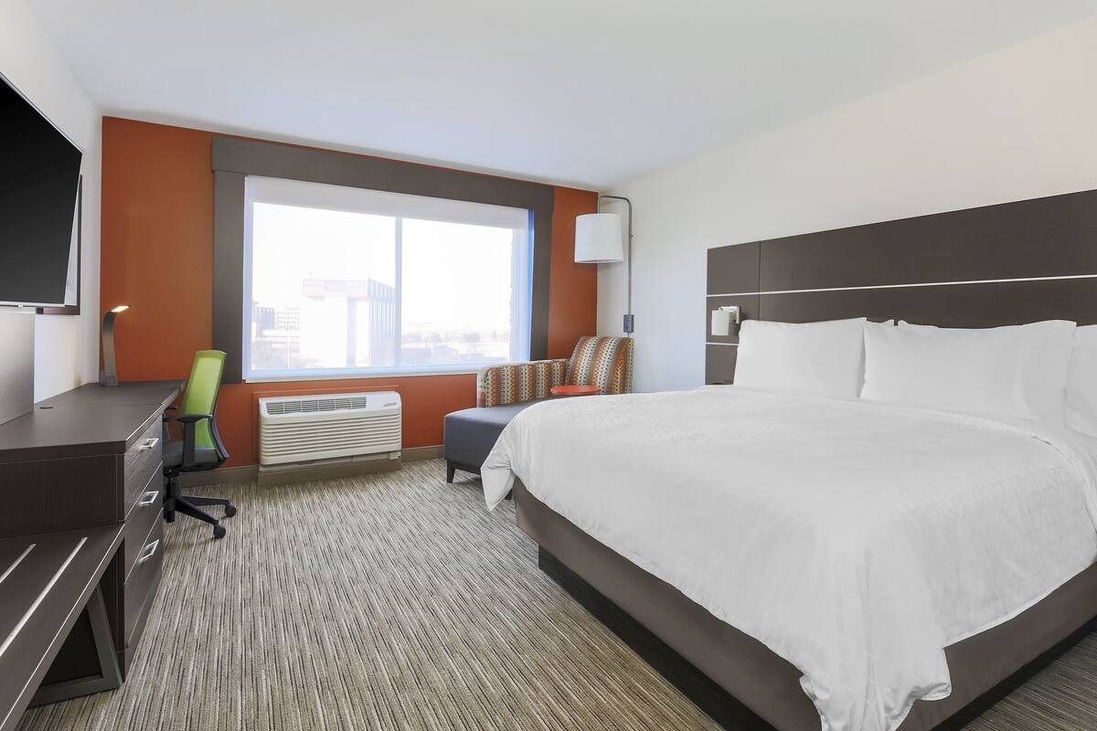 Habitación de hotel con una cama grande, un escritorio con cajones, una silla, ventana con vistas y un televisor montado en la pared.