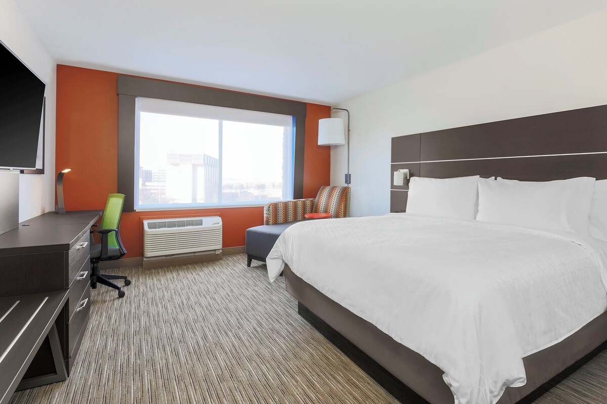 A hotel room with a large bed, a desk with drawers, a chair, a window, and a wall-mounted TV.