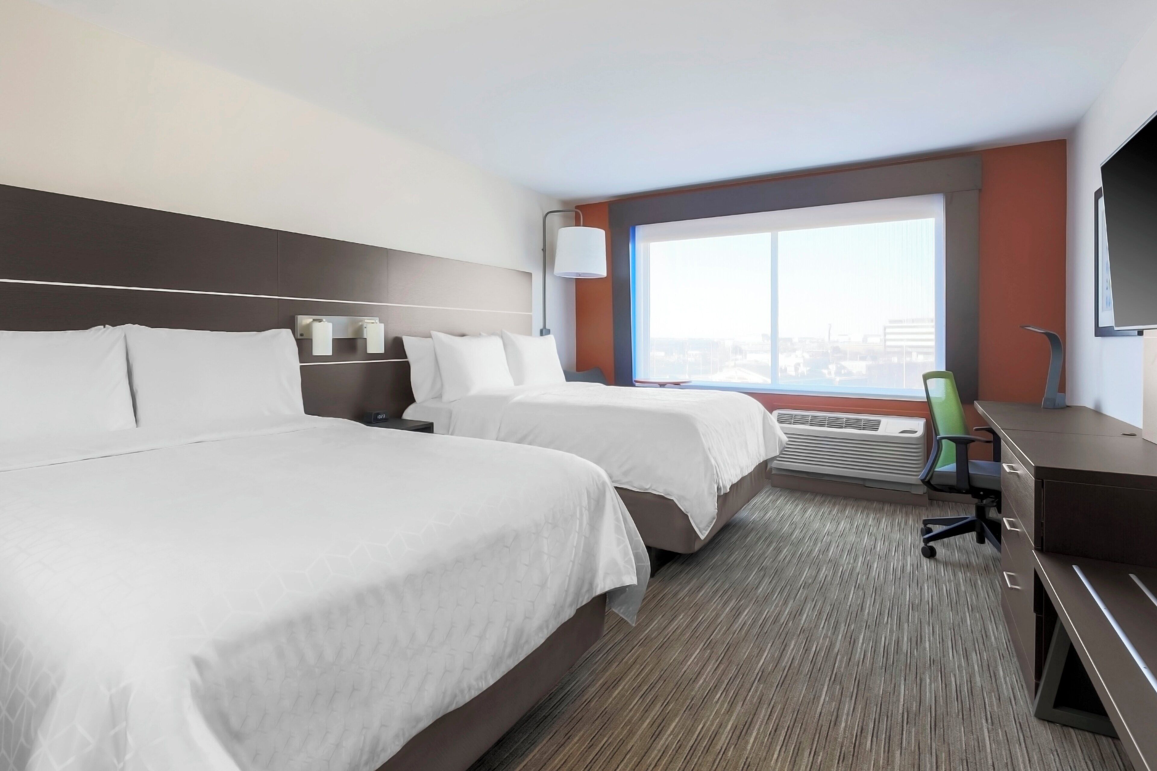A hotel room with two beds, a desk, a chair, a TV, a window with a city view, and a wall-mounted air conditioning unit.