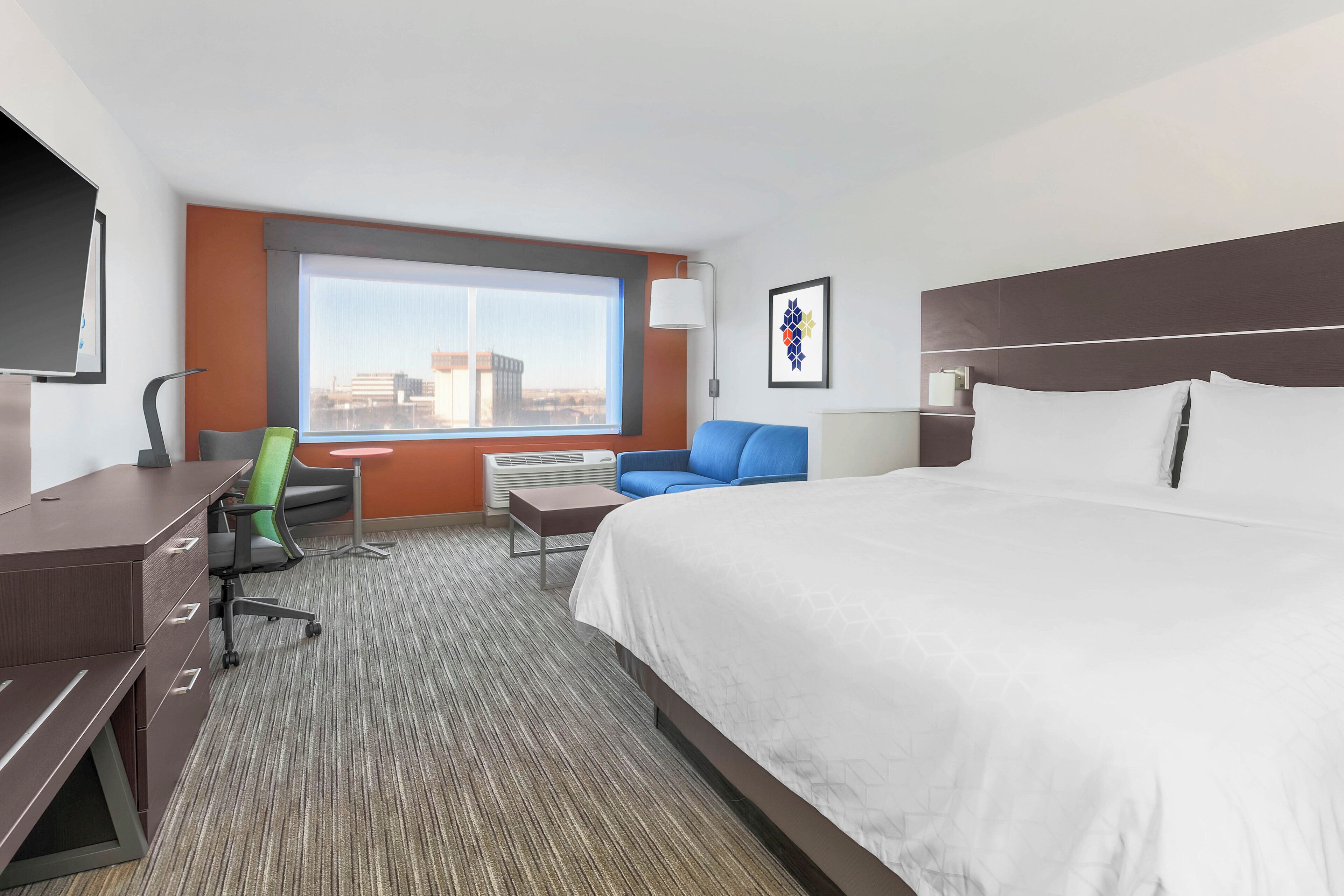 A hotel room with a large bed, a desk, a chair, a small table, a lamp, and a window with a city view.