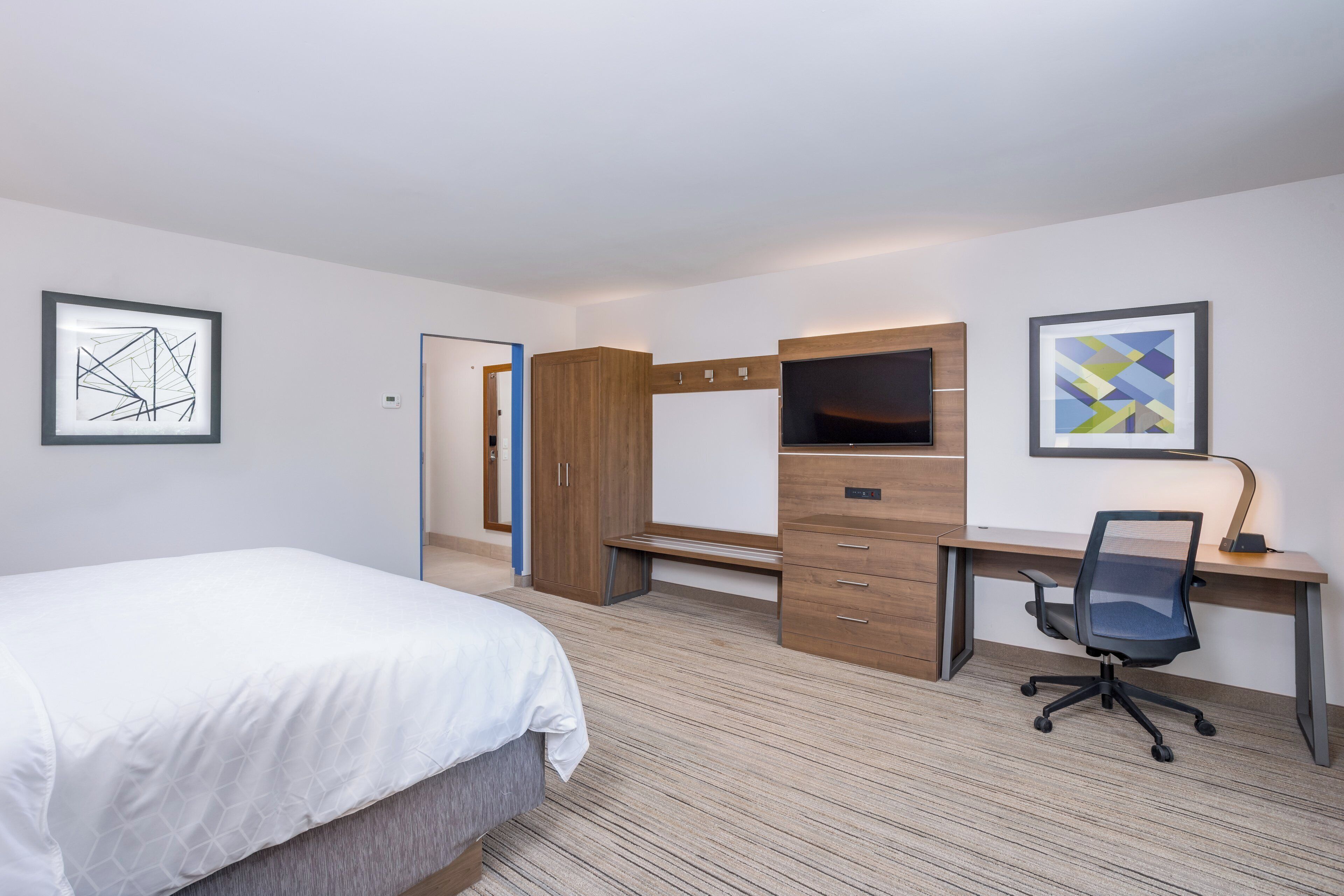 A hotel room with a bed, desk, chair, wardrobe, and artwork on the walls.