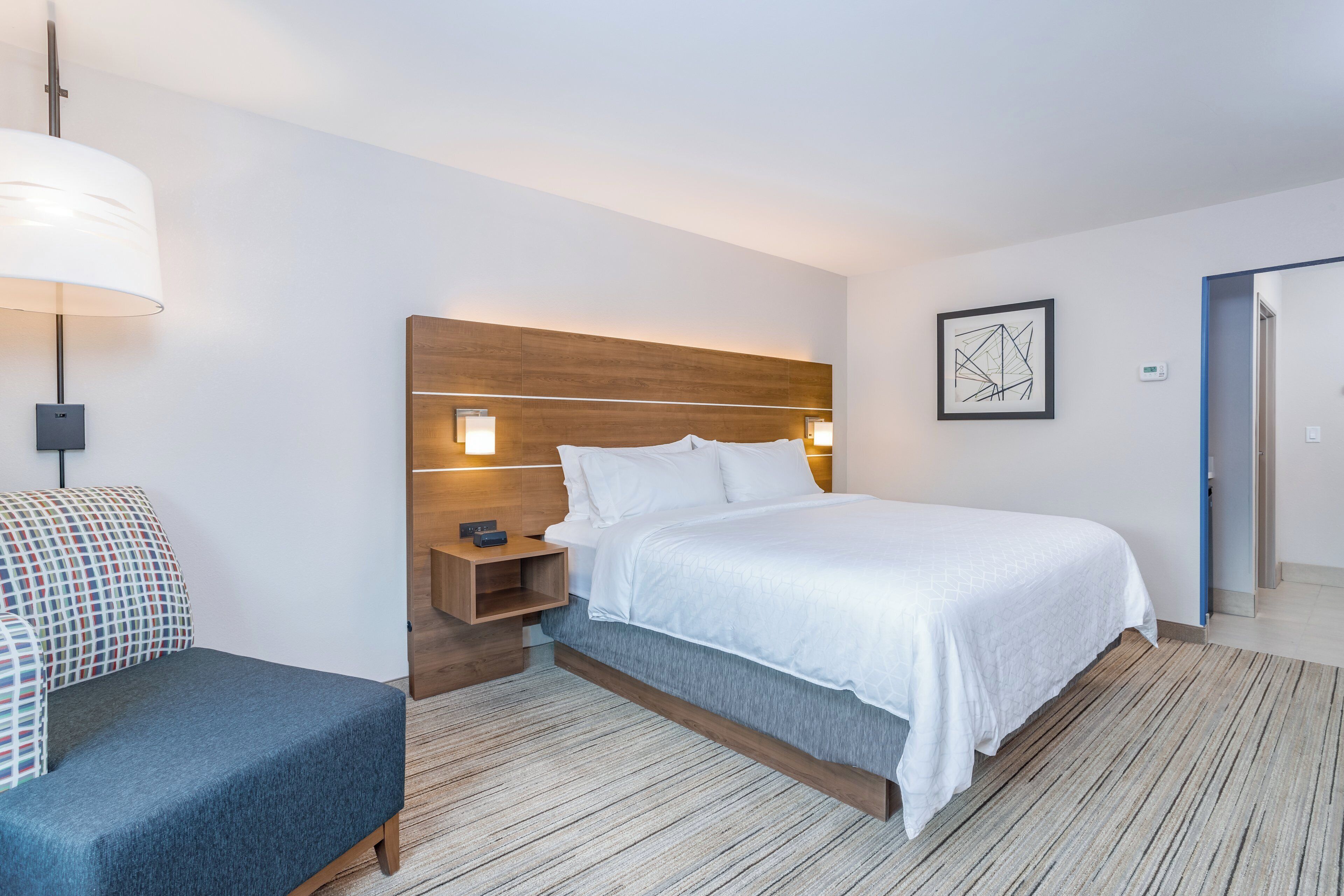A hotel room with a bed, bedside table, lamp, and a framed abstract artwork on the wall.