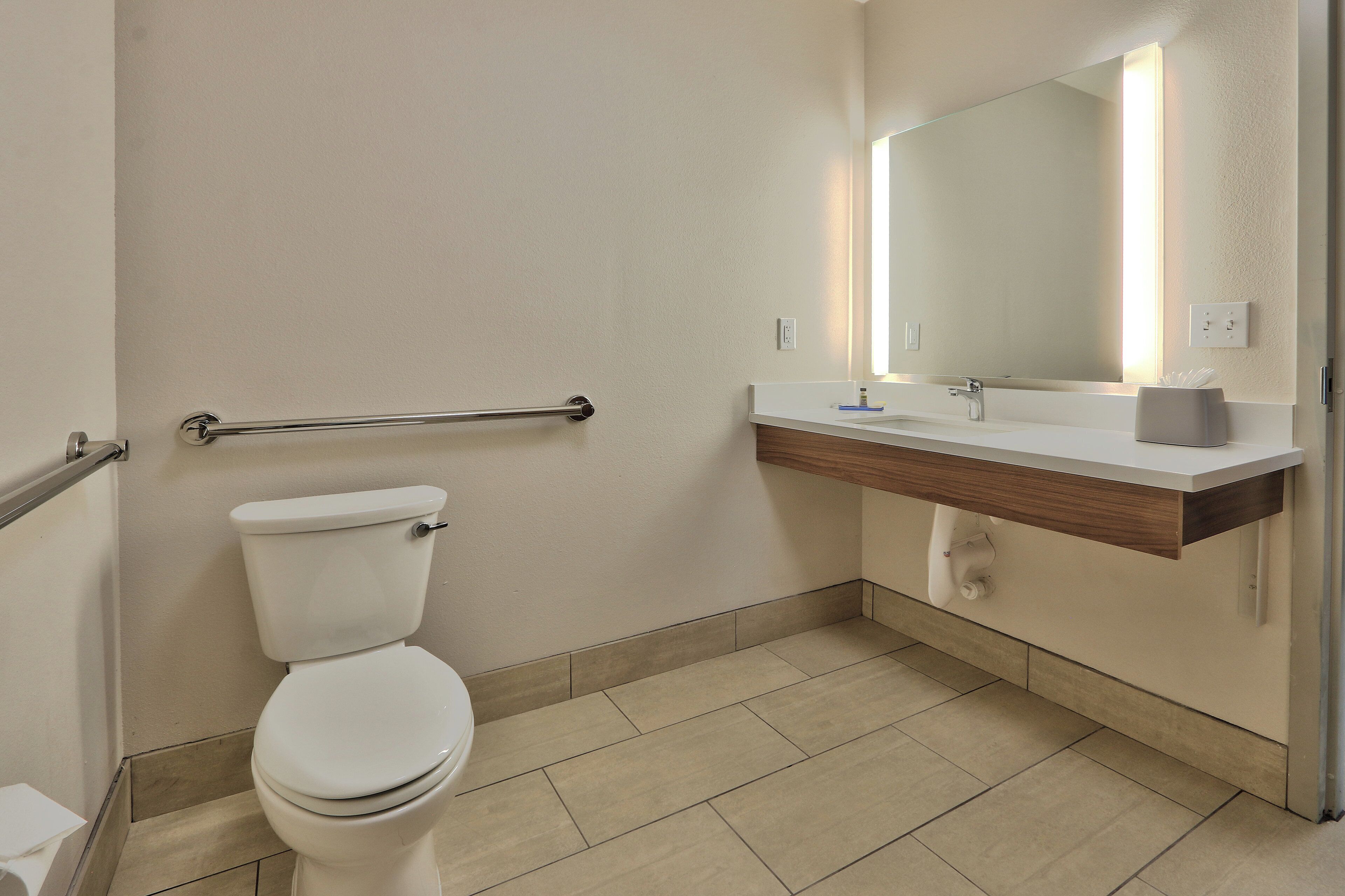 A standard hotel bathroom with a toilet, a sink, a mirror, and a handrail.