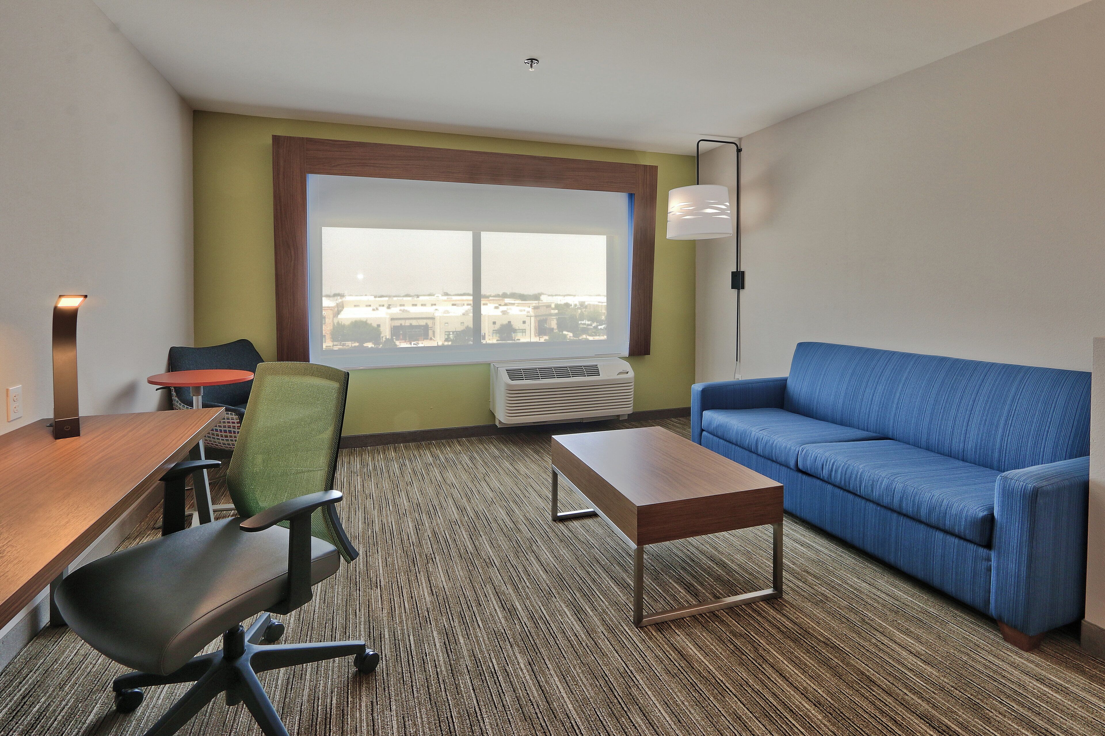 A hotel room with a large window, a blue sofa, a wooden desk, a green chair, and a standing lamp.
