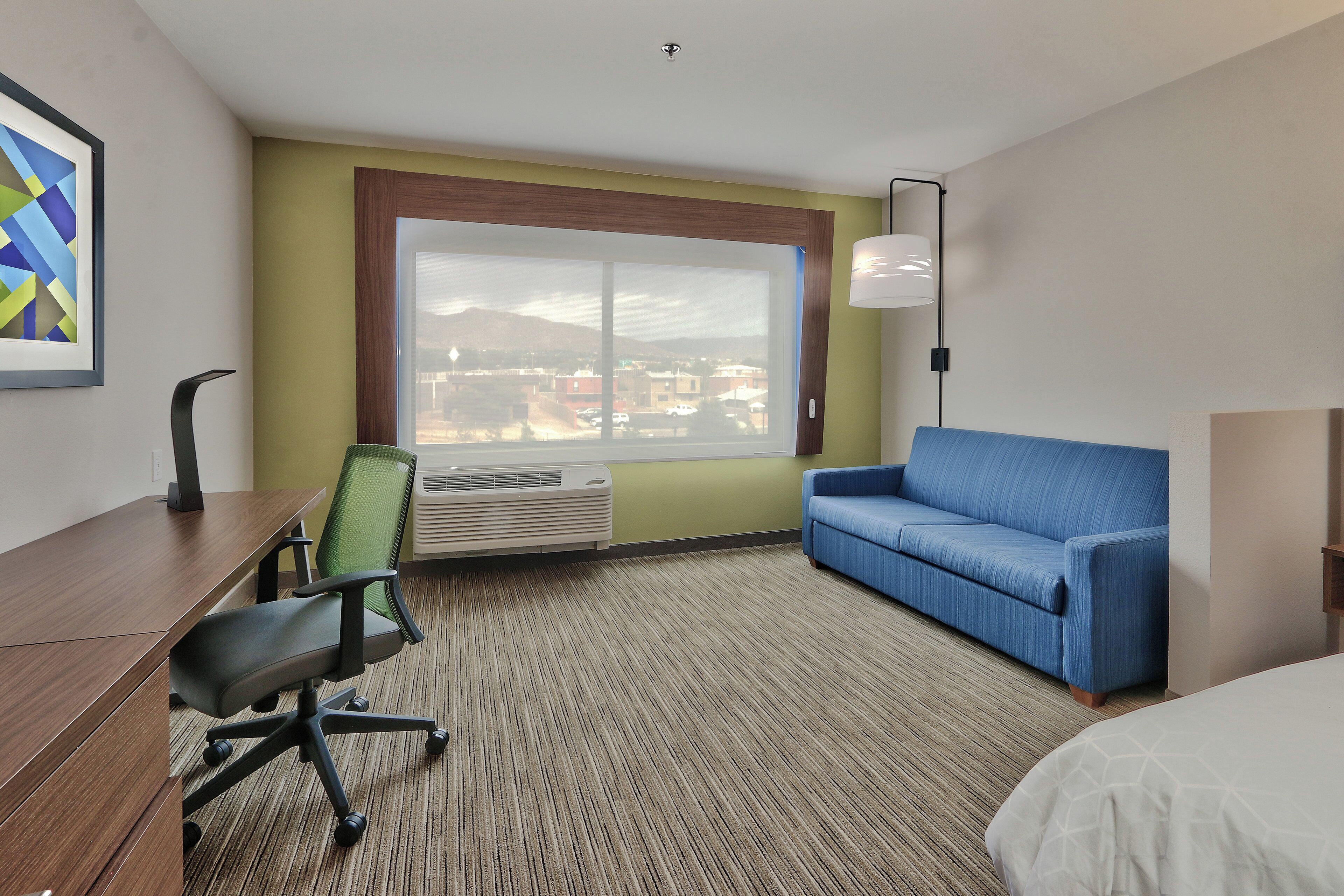 Habitación de hotel con escritorio, sofá azul, cama, ventana grande con vistas y un cuadro en la pared.