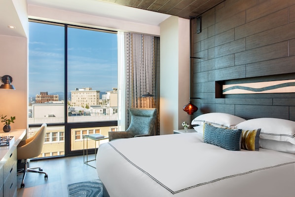 A modern hotel room with a large bed, a desk, and a chair. There is a view of the city through large windows.
