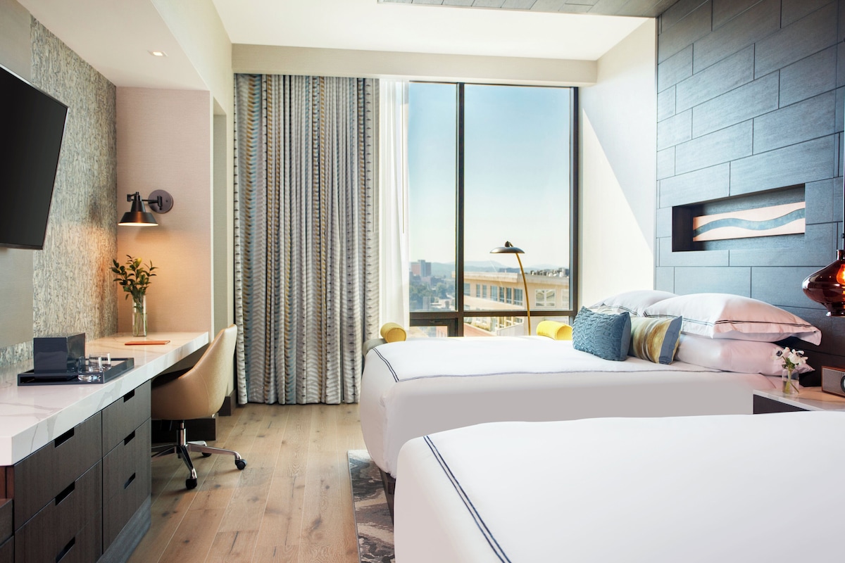 Habitación de hotel con escritorio, dos camas, televisor y un amplio ventanal con vistas a la ciudad.