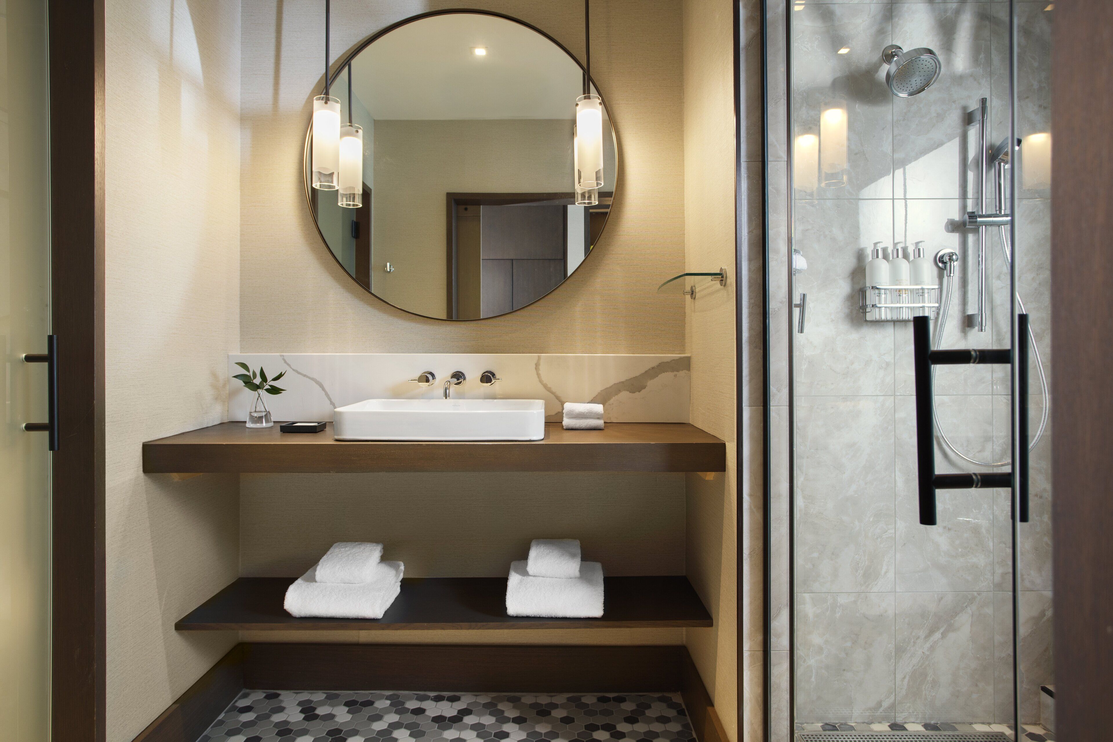 Une salle de bain moderne avec un miroir rond, un lavabo blanc et une douche séparée par des parois en verre.