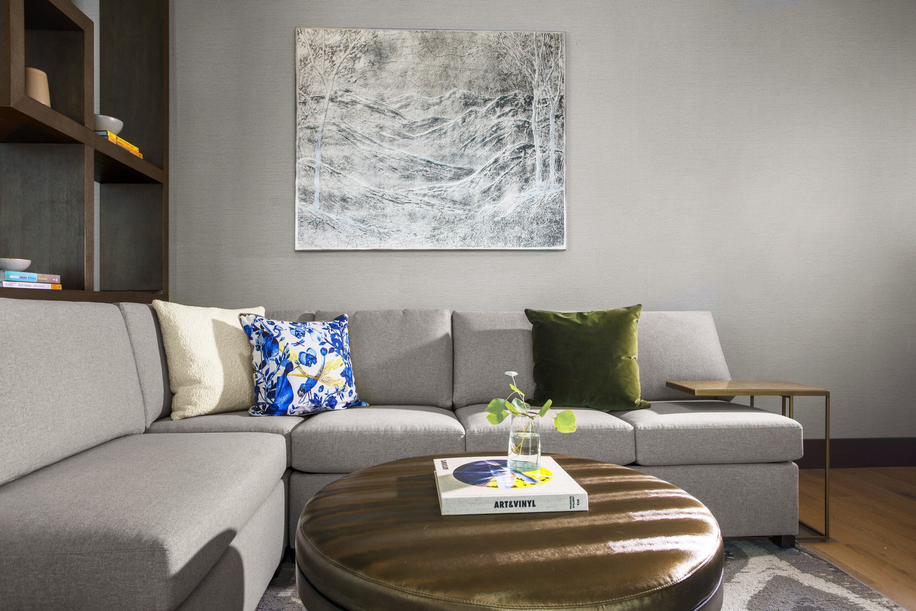 A modern living room with a grey sofa, a round leather ottoman, and a coffee table with a book and a vase.