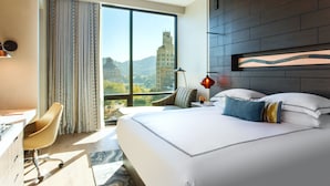 A modern hotel room with a large bed, a desk, and a chair. There is a view of a cityscape through the window.