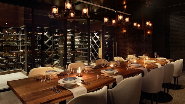 A long dining table set for a meal with white chairs, wine glasses, and a wine rack in the background.