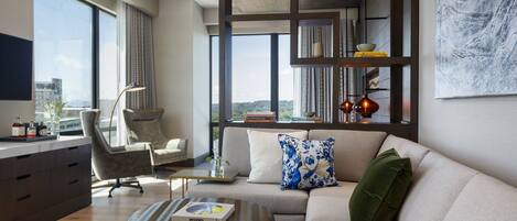 Una sala de estar moderna con sofá, sillón y mesa de centro. Hay una estantería con libros y objetos decorativos. Amplias ventanas ofrecen vistas de la ciudad.