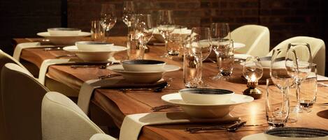 Una mesa de comedor dispuesta con platos, cuencos y cubiertos, rodeada de sillas, en una habitación con paredes de ladrillo y lámparas colgantes.