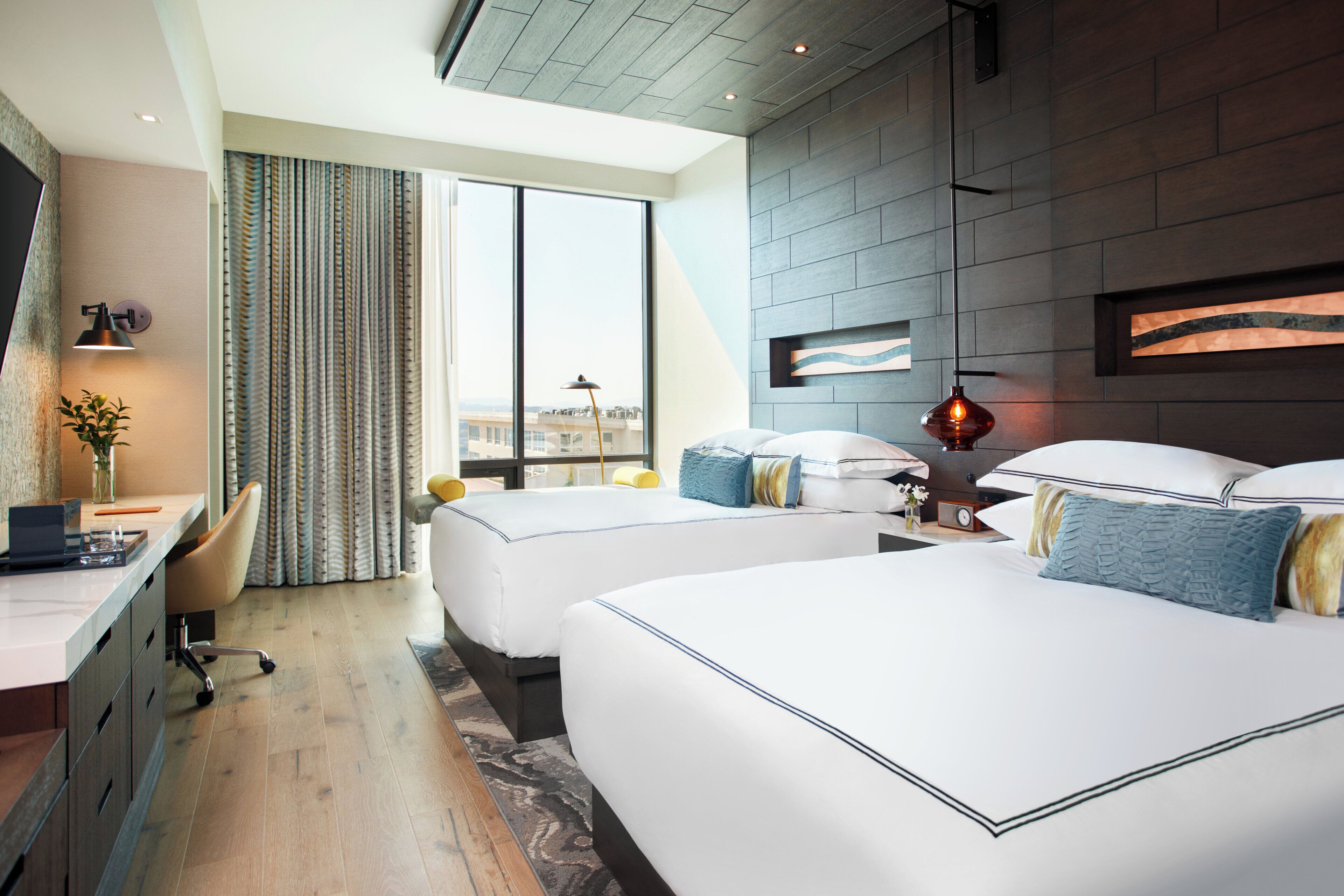 A hotel room with two beds, a desk, and a large window with curtains.