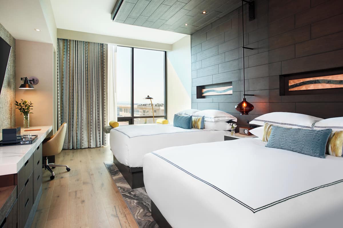 A hotel room with two beds, a desk, and a large window with curtains.