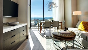 A modern hotel room with a large window, a flat-screen TV, a dining area, and a sofa with a lamp.