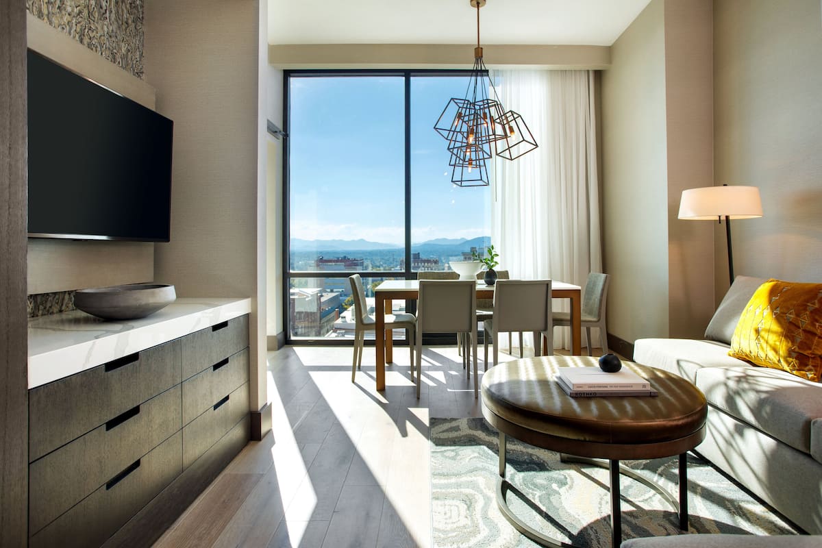 A modern hotel room with a large window, a flat-screen TV, a dining area, and a sofa with a lamp.