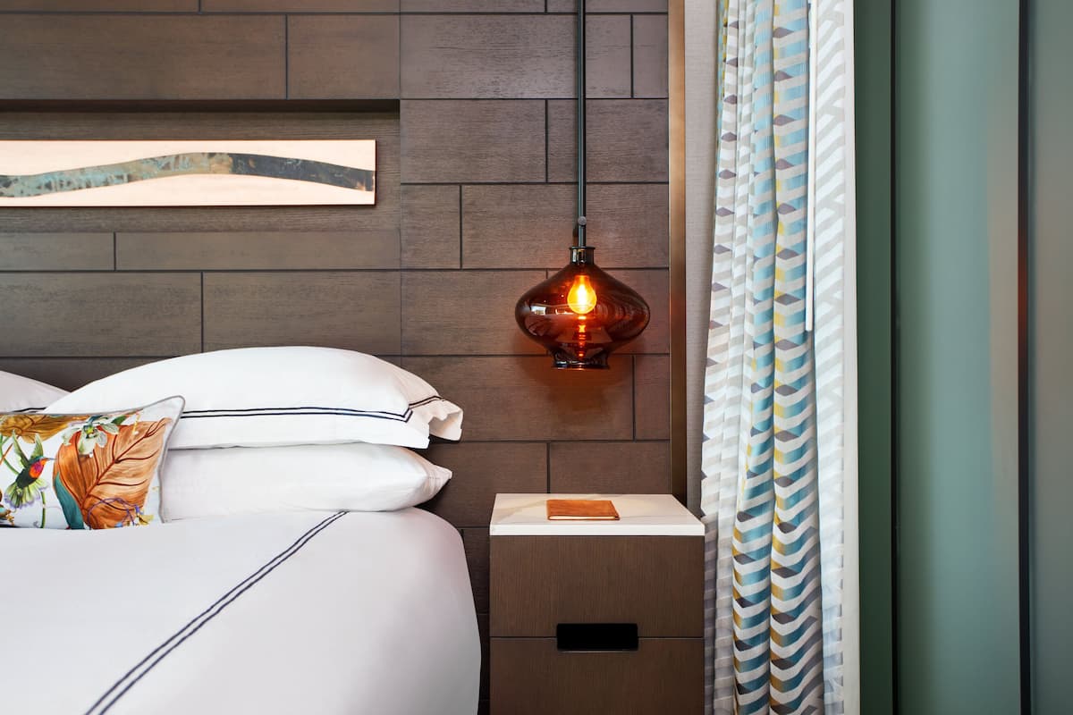 A neatly made bed with white linens and a decorative pillow, a bedside table with a book, a wall-mounted lamp, and a patterned curtain.